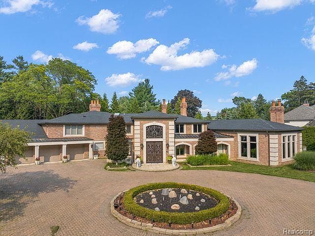 This is a front exterior view of a large, luxurious brick home. The house features a circular driveway with a decorative fountain in the center, manicured lawns, and mature trees. The architectural style is traditional with a grand entrance featuring an arched doorway and decorative ironwork.