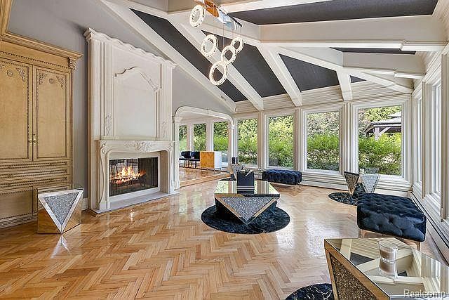 This is an interior shot of a luxurious living room featuring a vaulted ceiling with dark accents and exposed beams. The room is well-lit with natural light streaming through large windows, highlighting the herringbone wood flooring and a grand fireplace. The decor includes modern, geometric furniture and a built-in cabinet, creating an elegant and inviting space.