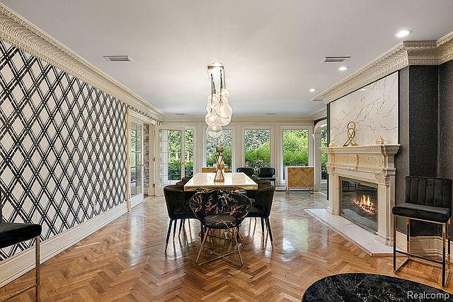 This is an interior shot of a dining room featuring herringbone wood floors, a long dining table with modern chairs, and a unique chandelier. The room has a fireplace and large windows that provide natural light, creating an elegant and inviting atmosphere. The walls are decorated with a geometric pattern, adding a touch of contemporary style.