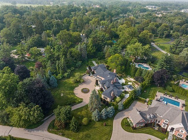 This aerial view showcases a sprawling estate with lush landscaping, mature trees, and multiple structures. The main house features a dark roof and manicured gardens, while a separate pool area and additional buildings add to the property's appeal. The overall impression is one of luxury and privacy.