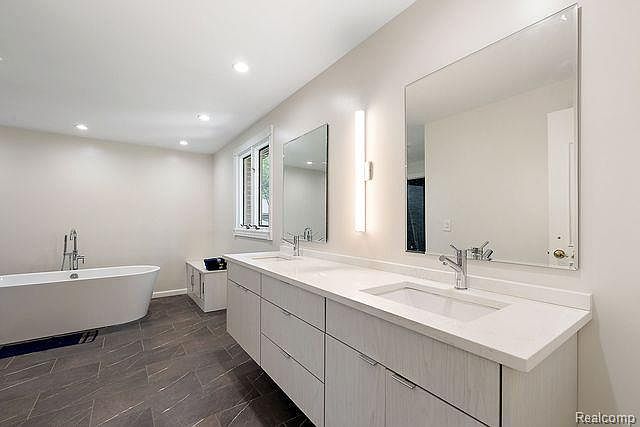 This is a primary bathroom featuring a double vanity with a white countertop and light wood cabinets. Two large mirrors hang above the sinks, and a freestanding bathtub is visible to the left. The floor is tiled with a dark gray material, and the overall style is modern and minimalist.