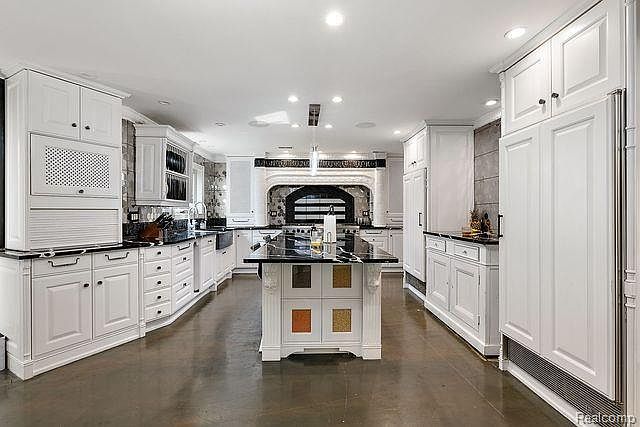 This is a spacious kitchen featuring white cabinetry, black countertops, and stainless steel appliances. A large island with a black countertop and decorative panels stands in the center of the room. The kitchen has a clean and elegant design, with a focus on functionality and style.