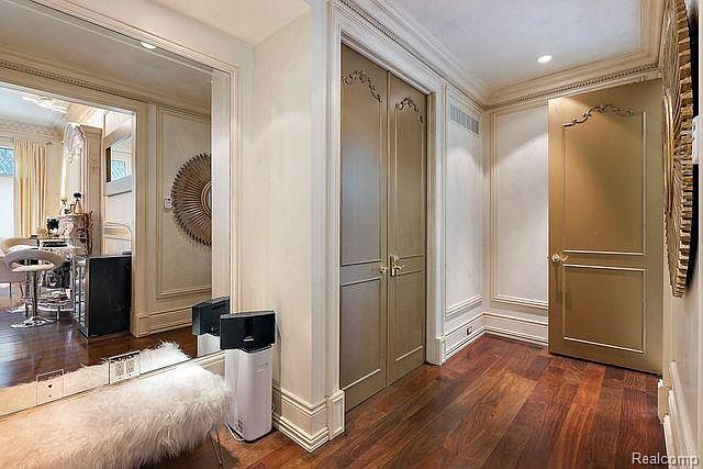 This interior shot showcases a luxurious hallway with rich hardwood flooring and ornate wall paneling. A mirrored bench with a faux fur cushion sits to the left, while a pair of paneled doors with decorative hardware are visible straight ahead. A gold-toned door adds a touch of elegance, and the overall impression is one of sophisticated design and high-end finishes.
