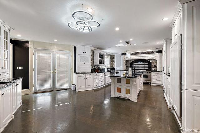 This is a spacious kitchen featuring white cabinetry, black countertops, and a large central island with a black countertop. The kitchen is well-lit with modern ceiling fixtures and recessed lighting. The dark wood flooring adds a touch of elegance to the space.