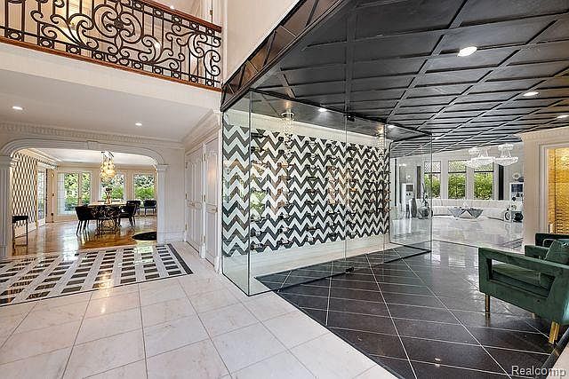 This interior shot showcases a grand hallway with a striking black and white patterned floor leading into a dining area. A modern, glass-enclosed wine cellar with a chevron pattern adds a unique architectural element. The space is well-lit and exudes a luxurious and contemporary feel.