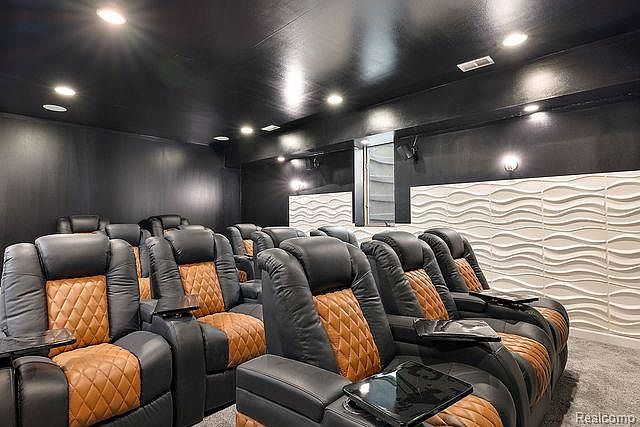 This is a home theater featuring multiple rows of black leather recliners with brown diamond-patterned accents. Each chair has a small table attached, and the room is painted in dark colors with recessed lighting to enhance the viewing experience. A textured white wall adds a modern touch to the space.