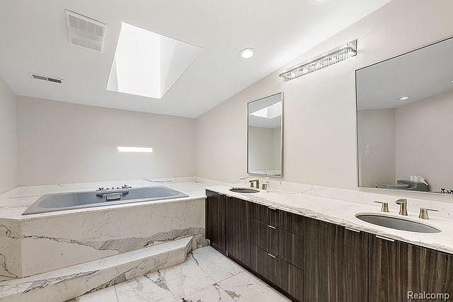 This is a primary bathroom featuring a large soaking tub with marble surround and steps, a double vanity with dark wood cabinets and marble countertop, and two large mirrors. The room is well-lit with a skylight and modern lighting fixture, creating a bright and luxurious atmosphere. The perspective is from the doorway, showcasing the entire space.