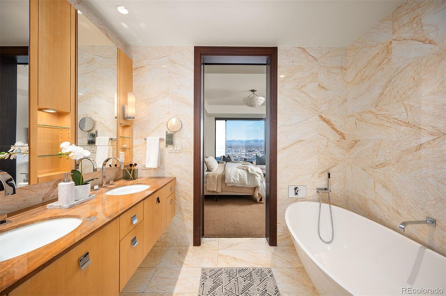This is a luxurious primary bathroom featuring a double vanity with light wood cabinetry and marble countertops. A large soaking tub sits adjacent to a doorway leading into the bedroom, offering a view of the bed and city skyline. The walls are covered in marble tiles, and the overall design is modern and elegant.