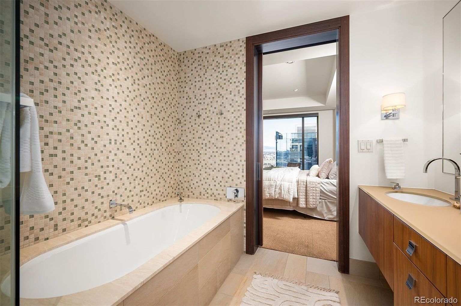 This is a primary bathroom featuring a modern design with a mosaic tiled wall above a soaking tub. The bathroom opens into a bedroom with a large window offering city views. A sleek vanity with a light-colored countertop and wood cabinetry is visible, enhancing the luxurious feel of the space.