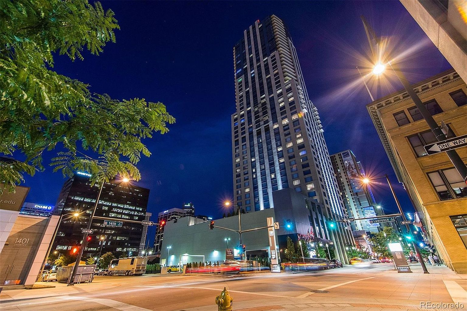 This image showcases a striking front view of a modern high-rise building at night, emphasizing its height and architectural details. The building features a sleek design with numerous illuminated windows, creating a visually appealing contrast against the dark sky. The surrounding street scene includes streetlights, traffic, and adjacent buildings, contributing to a vibrant urban atmosphere.