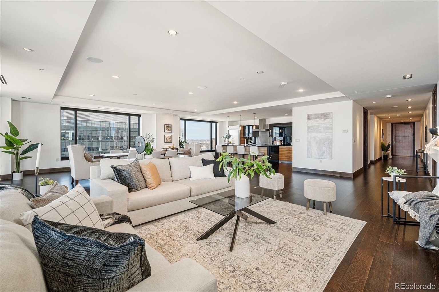 This is a spacious and modern living room featuring an open floor plan that seamlessly connects to the kitchen and dining area. The room is furnished with a large sectional sofa, a glass coffee table, and stylish accent chairs. Dark hardwood floors and large windows offering city views add to the luxurious feel of the space.