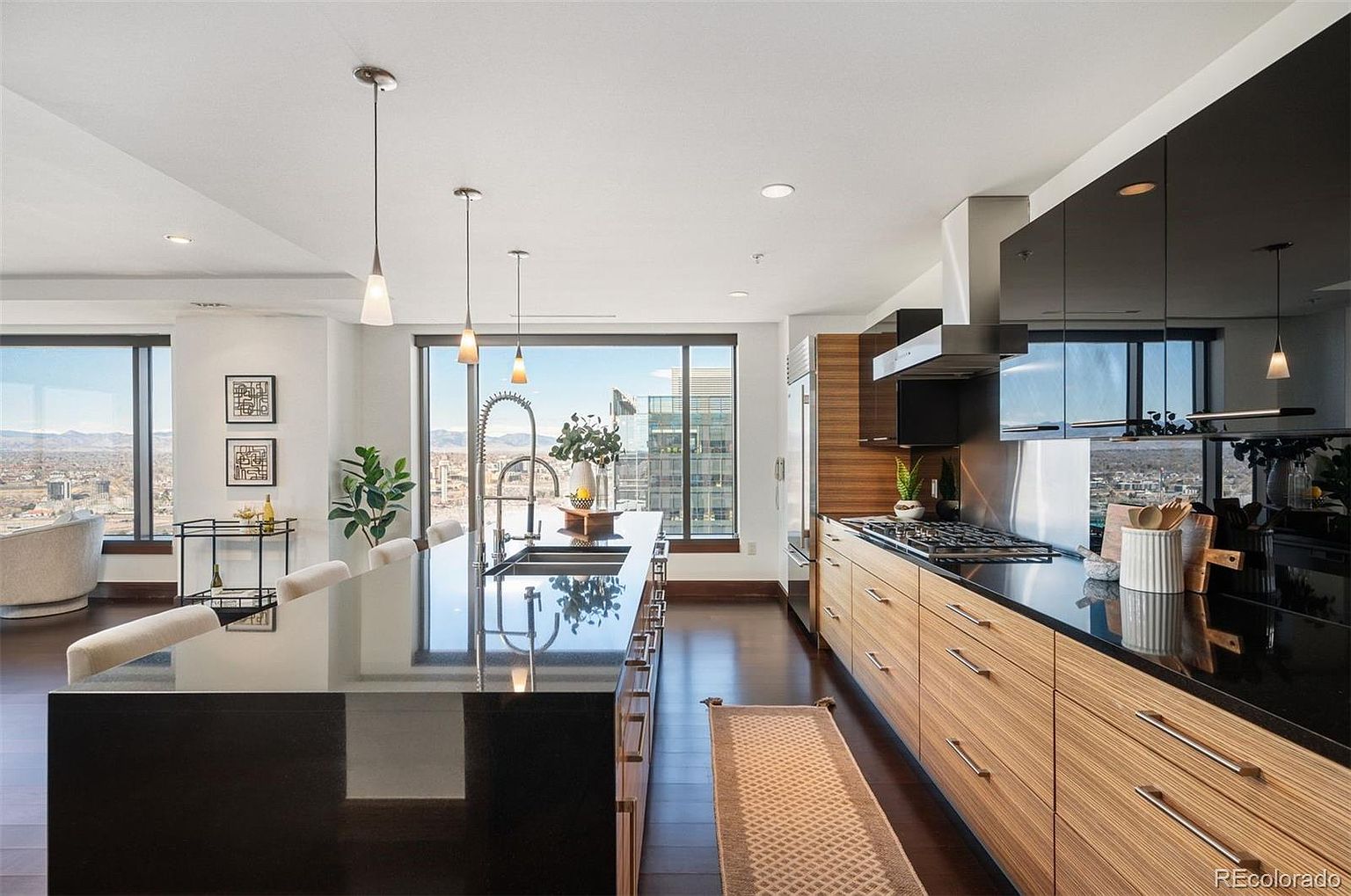 This is a modern kitchen with sleek, dark cabinetry contrasted by light wood drawers and cabinets. A large island with a black countertop dominates the space, complemented by pendant lighting. Large windows offer a stunning city view, enhancing the kitchen's luxurious and contemporary feel.