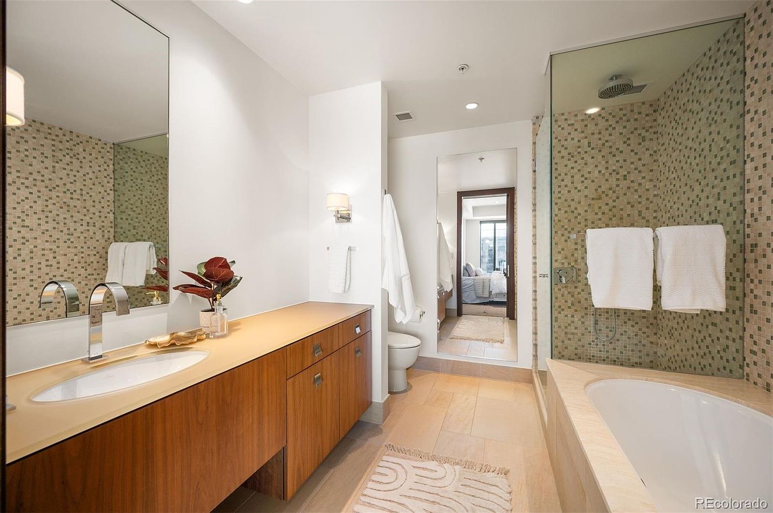 This is a well-lit primary bathroom featuring a long wooden vanity with a white sink, a large mirror, and mosaic tile accents on the walls. A glass-enclosed shower and bathtub combination is visible on the right, with white towels neatly hung. The flooring is light-colored tile, and a small rug adds a touch of warmth to the space.