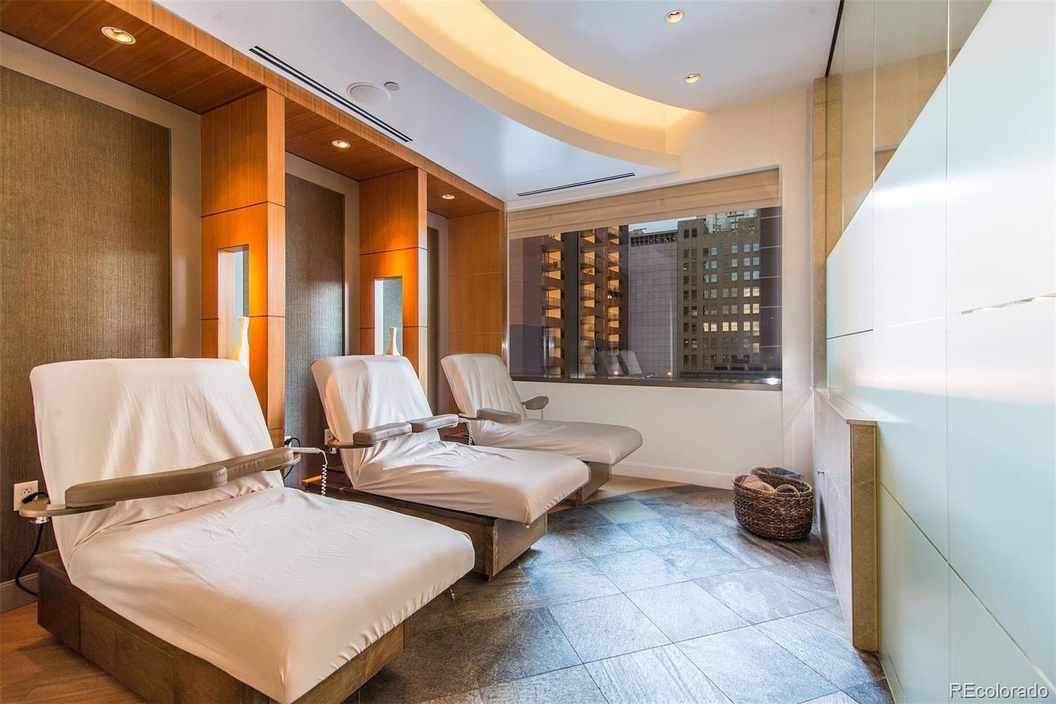 This interior shot showcases a relaxation or spa area, featuring three cushioned lounge chairs draped with white covers. The room is illuminated by recessed lighting and natural light filtering through a window with a city view. The flooring is tiled, and the overall ambiance is serene and modern, suggesting a high-end amenity space.