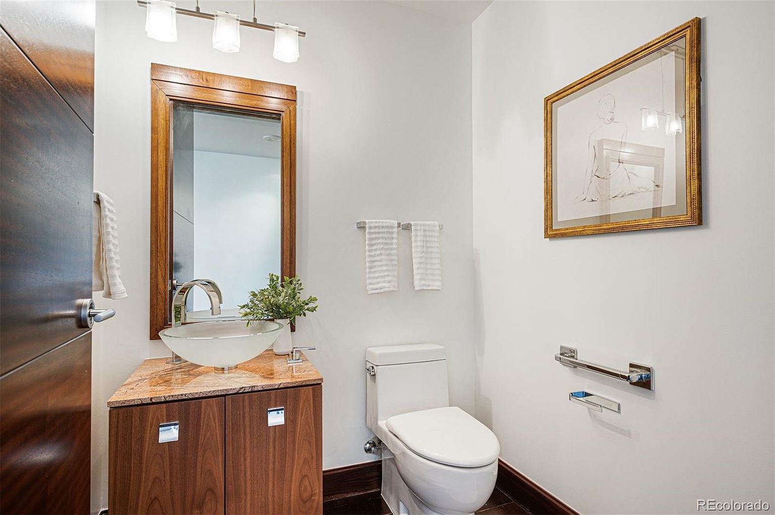This is a well-lit guest bathroom featuring a modern vanity with a vessel sink and a wood-framed mirror. A toilet is positioned next to a towel rack, and framed artwork hangs on the wall. The room has a clean and contemporary aesthetic, with a focus on functionality and style.