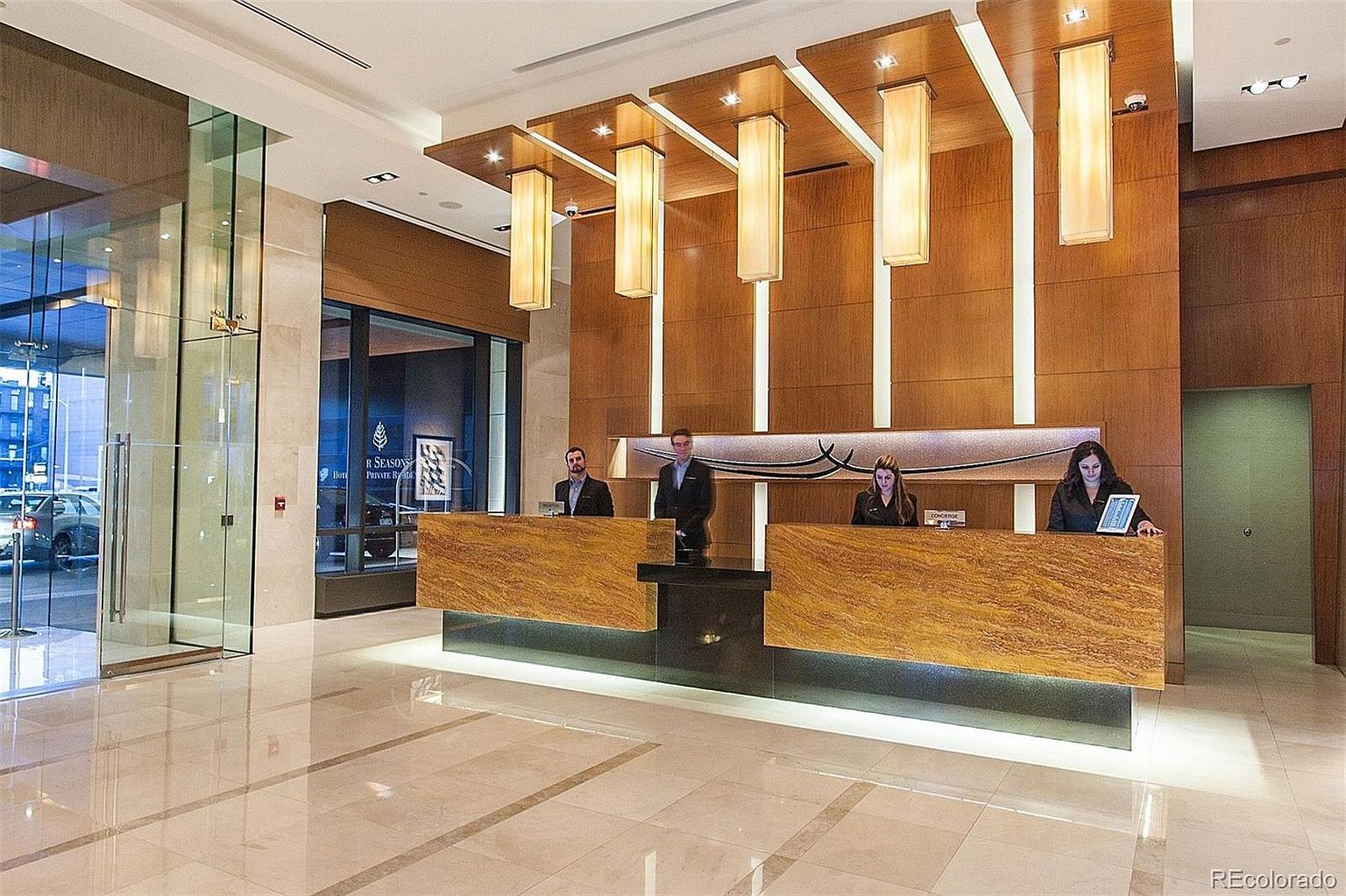 This is an interior shot of a hotel lobby featuring a reception desk with three staff members. The lobby has a modern design with wood paneling, contemporary lighting fixtures, and a polished marble floor. The overall impression is clean, upscale, and inviting.