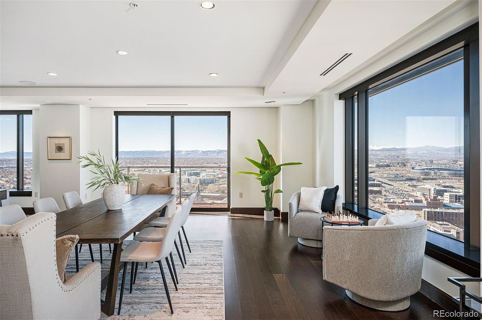 This interior shot showcases a modern dining room with a long wooden table surrounded by upholstered chairs. Large windows offer expansive city views, enhancing the room's bright and airy atmosphere. The dark hardwood floors contrast with the light walls and furniture, creating a sophisticated and inviting space.