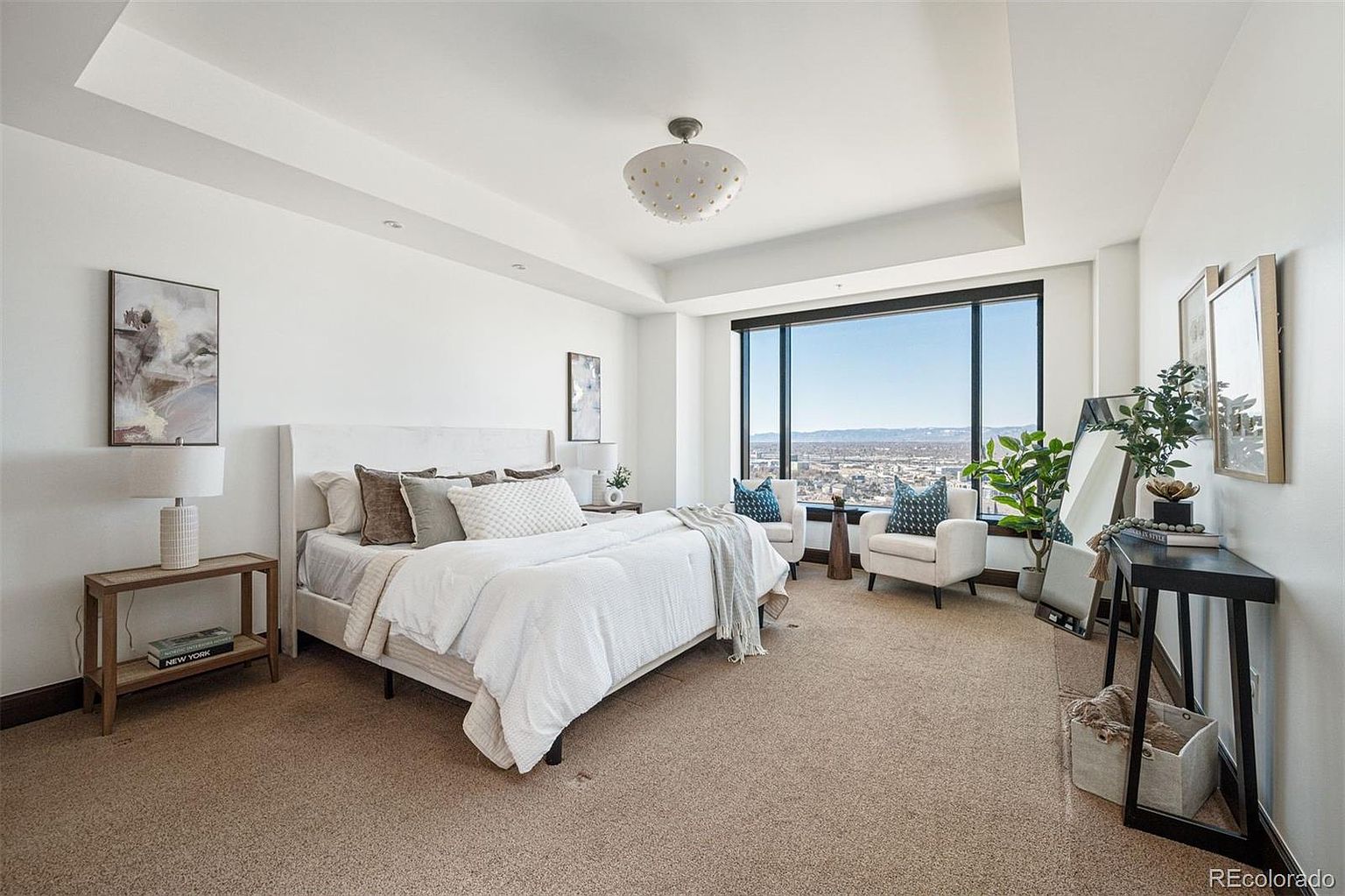 This is a bright and spacious primary bedroom featuring a large window with a city view. The room is decorated in neutral tones with a white bed, soft linens, and comfortable seating. A side table with a lamp and artwork adds a touch of elegance, while a console table with decorative items and a mirror completes the look.