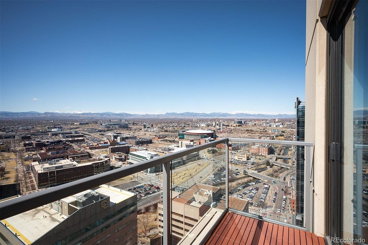 This image showcases a balcony view from a high-rise building, offering a panoramic cityscape under a clear blue sky. The balcony features a modern glass railing and a wooden deck, providing an inviting outdoor space. The view encompasses a sprawling urban landscape with distant mountains, highlighting the property's prime location and stunning vistas.