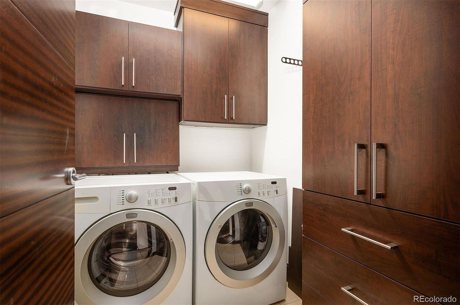 This is a well-organized laundry room featuring a front-loading washer and dryer set. Dark wood cabinets with sleek, silver hardware provide ample storage above and to the side of the appliances. The room appears modern and functional, ideal for efficient laundry management.