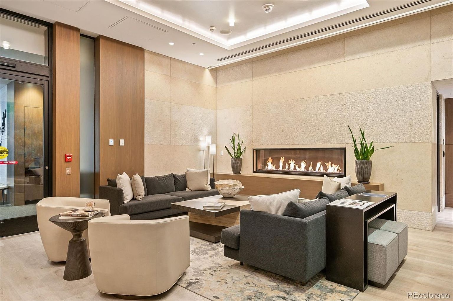 This is an interior shot of a modern living room featuring a linear fireplace, a gray sectional sofa with neutral pillows, and two cream-colored armchairs. The room is decorated with plants and a contemporary coffee table, and the walls are clad in a textured, light-colored material. The overall impression is one of sophisticated comfort and warmth.