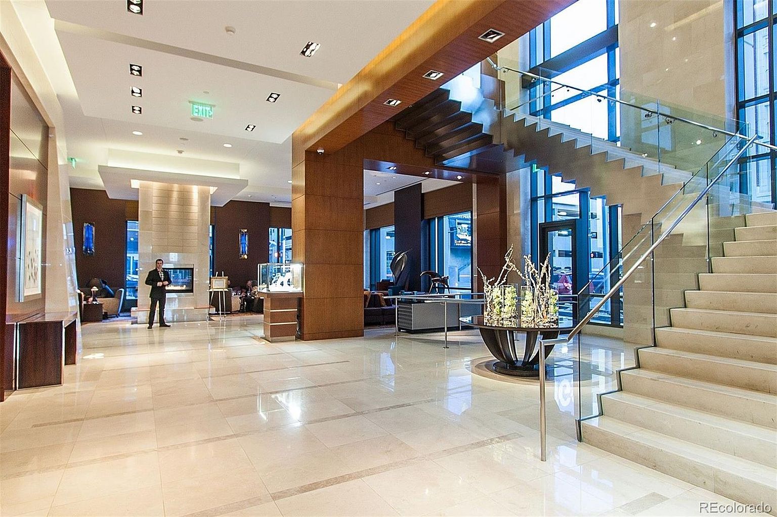 This is an interior shot of a luxurious lobby or hallway featuring a grand staircase with glass railings. The space is well-lit with a combination of natural light from large windows and recessed lighting. The flooring is a polished tile, and there are decorative elements such as a table with a floral arrangement and wood paneling that add to the upscale ambiance.