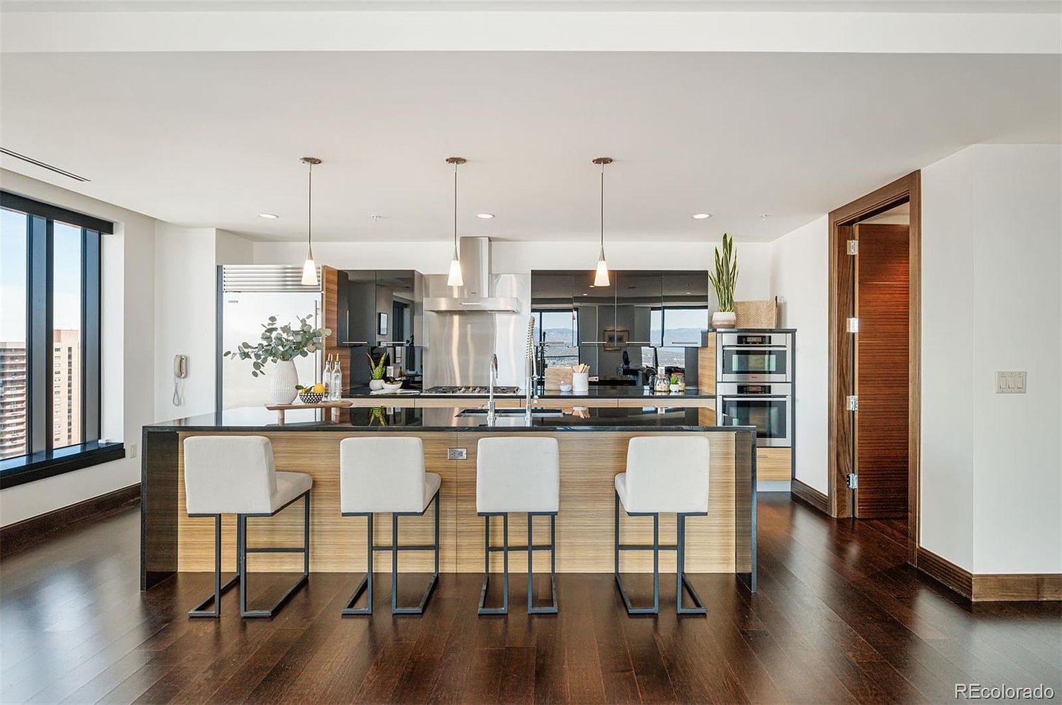 This is a modern kitchen featuring a large island with seating, dark countertops, and light wood cabinetry. Stainless steel appliances and pendant lighting add to the contemporary feel. A large window provides natural light and a view, while a doorway leads to another room, enhancing the open and airy atmosphere.