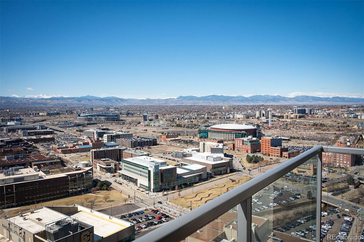 This aerial view showcases a sprawling cityscape under a clear blue sky, with distant mountains providing a scenic backdrop. The image captures a mix of modern buildings, stadiums, and parking lots, offering a sense of urban density and activity. The perspective is from a high vantage point, possibly a balcony or rooftop, providing a comprehensive overview of the area.