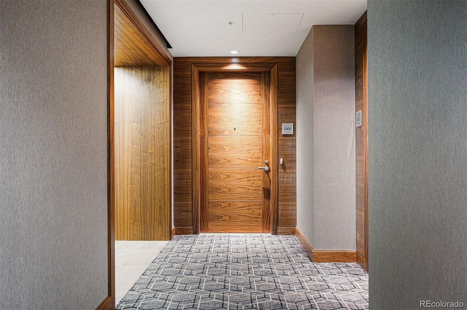 This image showcases a well-lit hallway with a sophisticated design. The hallway features a patterned carpet, wood-paneled walls, and a prominent wooden door with the number '2300' displayed. The overall impression is one of luxury and attention to detail, making it an appealing feature for a real estate listing.