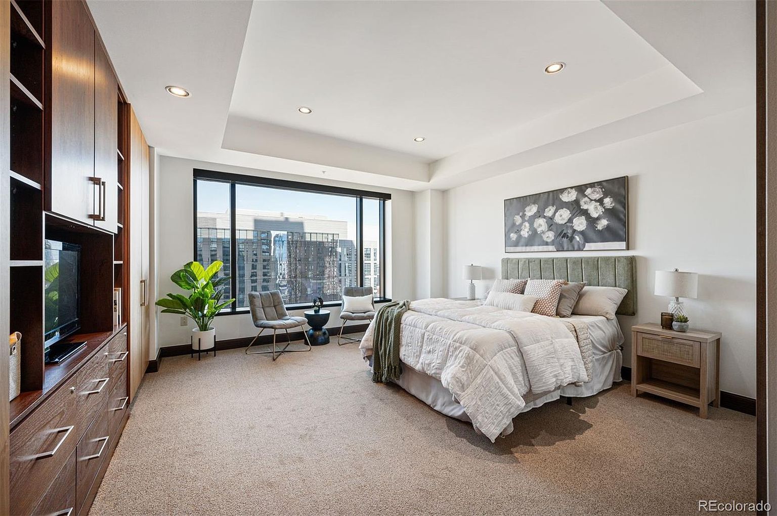 This is a well-lit primary bedroom featuring a large window with city views, a neutral color palette, and carpeted floors. The room includes a bed with a padded headboard, a nightstand with a lamp, and built-in wooden cabinetry with a television. Two chairs and a small table are positioned near the window, creating a cozy seating area.