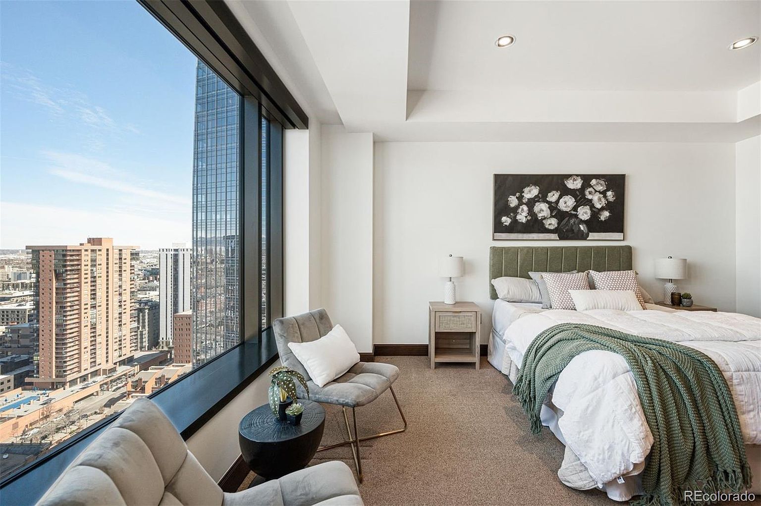 This is a primary bedroom featuring a large window with a city view. The room is decorated in neutral tones with a bed, nightstands, lamps, and seating area. The overall impression is modern and luxurious, with an emphasis on the view.