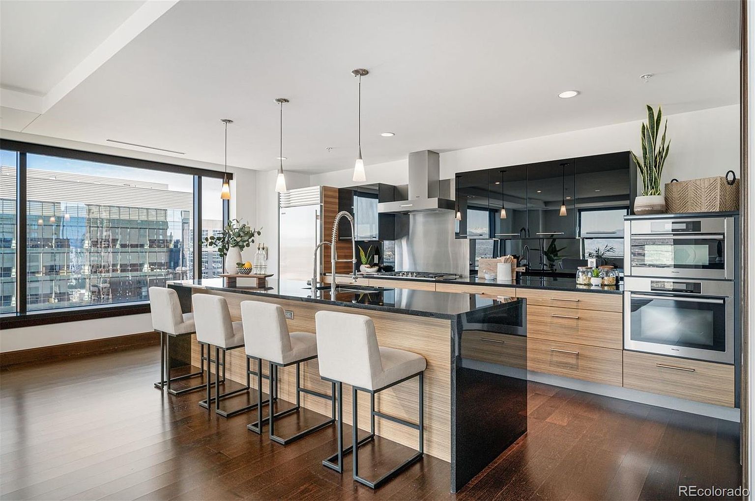 This is a modern kitchen featuring a large island with bar seating, sleek black cabinetry, and stainless steel appliances. The kitchen has a contemporary design with wood-grain accents and a large window offering city views. The dark hardwood floors add warmth and sophistication to the space.