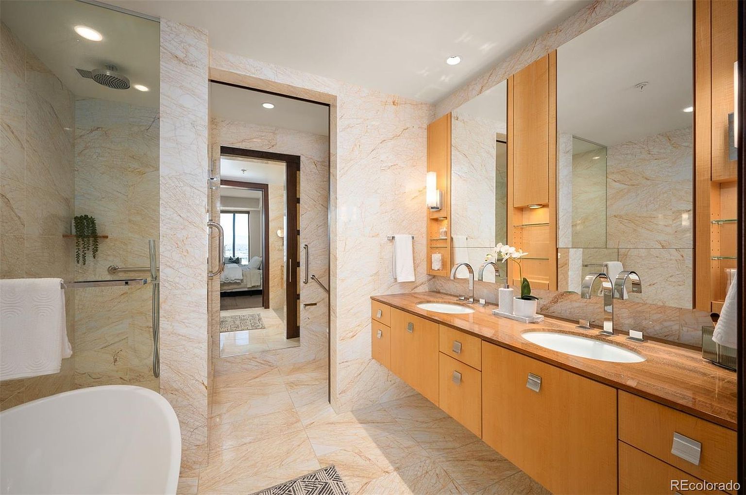 This is a luxurious primary bathroom featuring marble walls and flooring, a double vanity with wood cabinetry and a light-colored countertop, and a large mirror. The bathroom also includes a modern bathtub and a glass-enclosed shower. The overall impression is one of elegance and sophistication.
