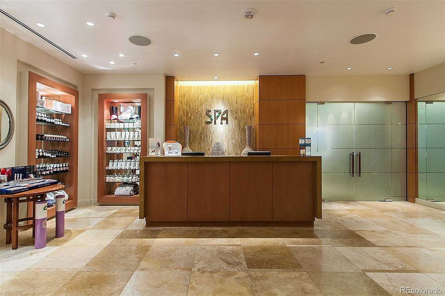 This image showcases the interior of a spa reception area. The focal point is a wooden reception desk with the word "SPA" displayed on the wall behind it. Display shelves filled with various products are visible on the left, and frosted glass doors are on the right, suggesting a tranquil and luxurious environment.