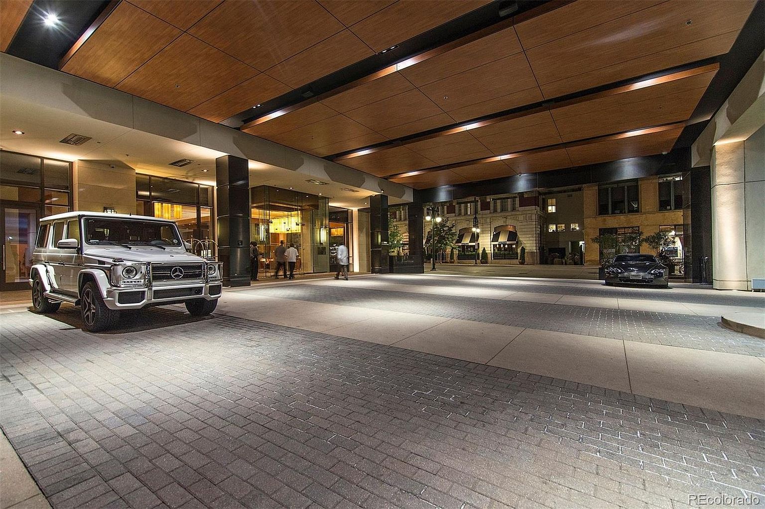 This image showcases the grand entryway of a building, featuring a covered driveway with a wooden ceiling and recessed lighting. A white Mercedes G-Wagon is parked prominently on the left, while a black car is visible further in the background. The entryway leads to glass doors and a well-lit interior, suggesting a luxurious and welcoming atmosphere.