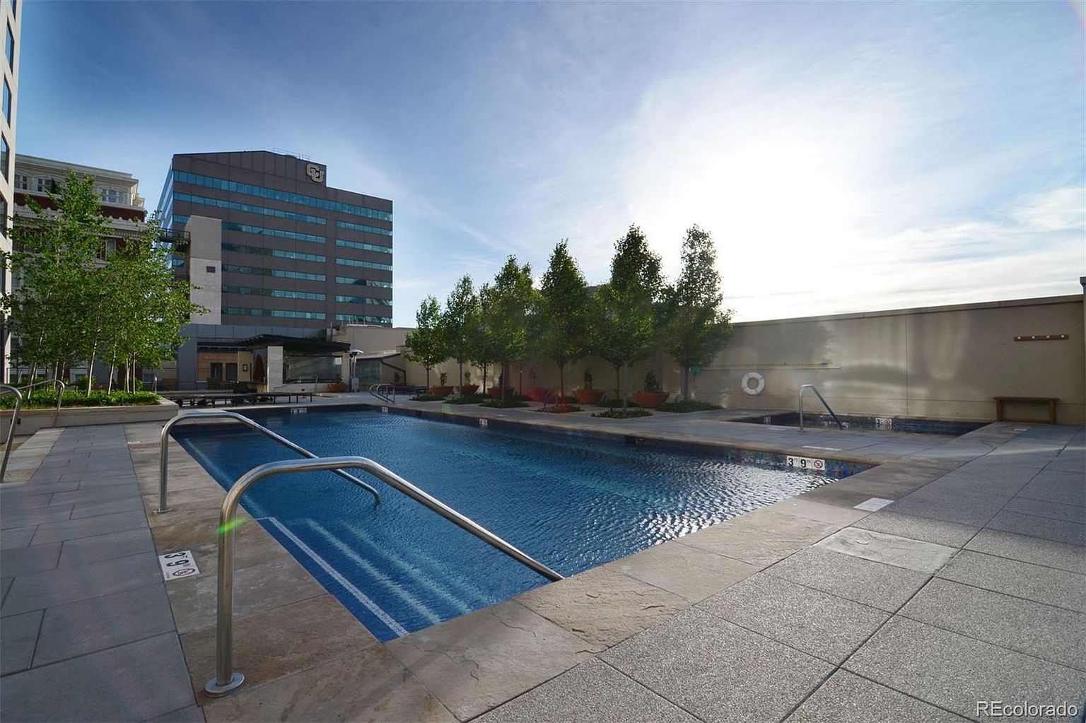 This image showcases a well-maintained outdoor pool and spa area, likely part of a condominium or apartment complex. The pool features clear blue water and is surrounded by a paved deck area with trees and a building in the background. The scene conveys a sense of relaxation and luxury, highlighting the property's amenities.