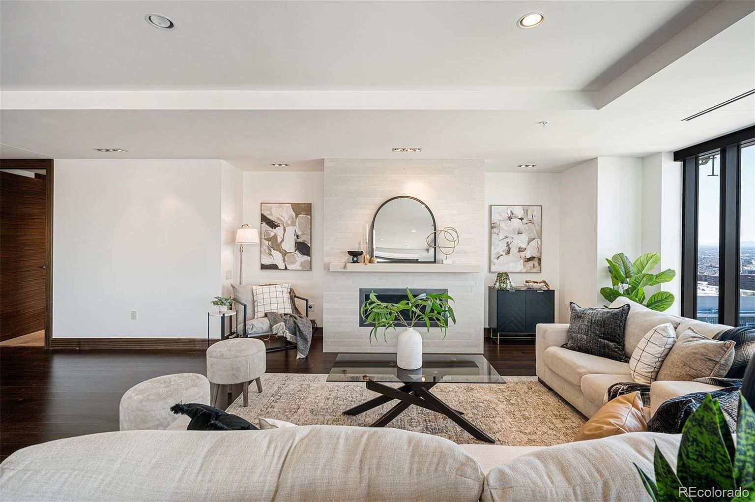This is a well-lit living room featuring a modern fireplace with a decorative mirror above it. The room is furnished with comfortable sofas, stylish ottomans, and a glass-topped coffee table, all arranged on a patterned area rug. Large windows offer a city view, enhancing the room's appeal.