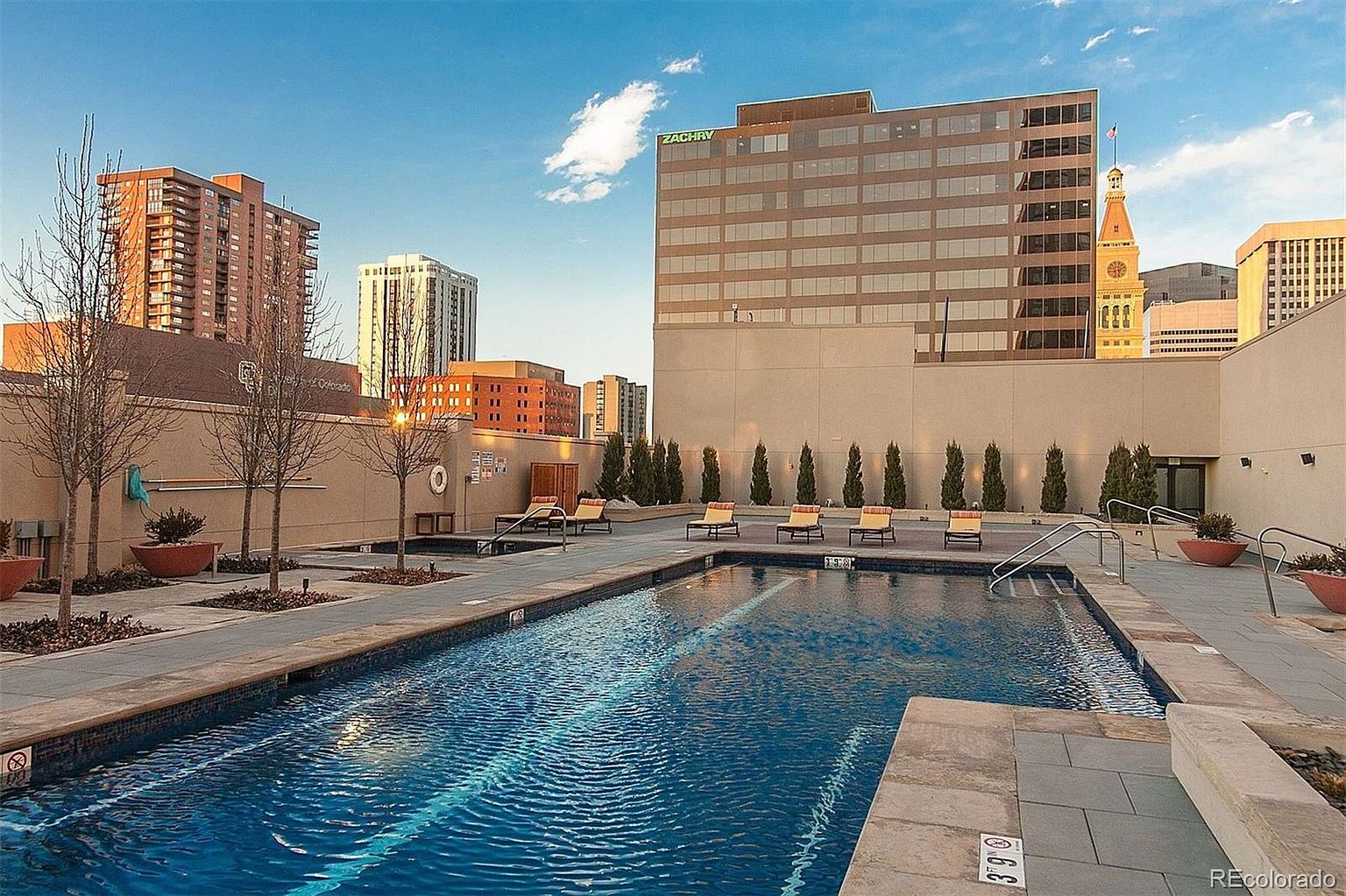 This image showcases a luxurious outdoor pool area in an urban setting. The pool is surrounded by a paved deck with lounge chairs and manicured greenery. Tall buildings rise in the background, creating a sophisticated and upscale atmosphere, perfect for relaxation and entertainment.