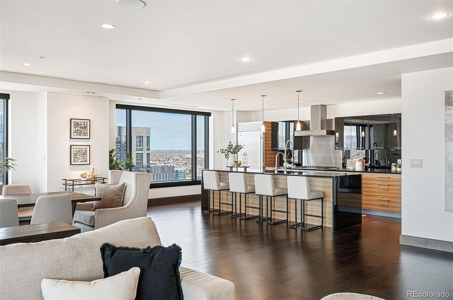 This is an interior shot of a modern living room and kitchen area. The space features dark hardwood floors, a large kitchen island with bar seating, and floor-to-ceiling windows offering a city view. The living area includes a comfortable sofa and chairs, creating an open and inviting atmosphere.
