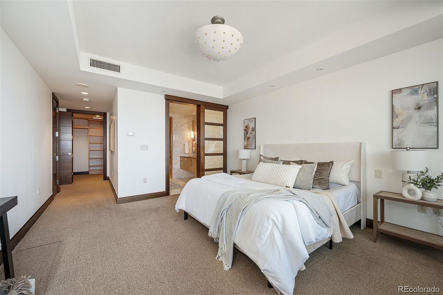 This is a primary bedroom featuring a neutral color palette with white walls and beige carpet. The room includes a bed with a white headboard, several decorative pillows, and a light-colored throw blanket. There is a sliding door leading to the bathroom and a walk-in closet, enhancing the room's functionality and appeal.