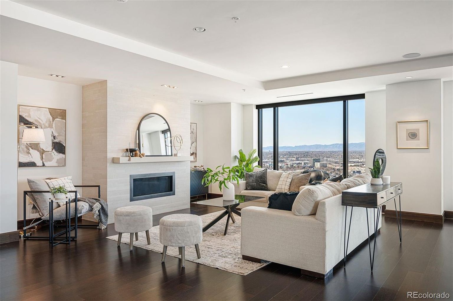 This is a bright and modern living room featuring dark hardwood floors, a neutral-toned sofa, and a fireplace with a decorative mirror above. Large windows offer a panoramic city view, enhancing the room's spacious feel. The room is tastefully decorated with plants, artwork, and stylish furniture, creating an inviting and sophisticated atmosphere.