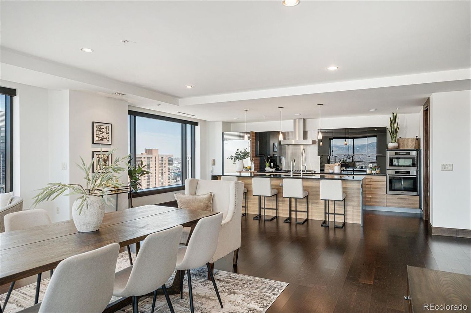 This is an open-concept kitchen and dining area featuring dark hardwood floors and modern design elements. The kitchen boasts stainless steel appliances, a large island with bar seating, and sleek cabinetry. The dining area includes a long wooden table with upholstered chairs, complemented by a stylish rug and large windows offering city views, creating a sophisticated and inviting atmosphere.