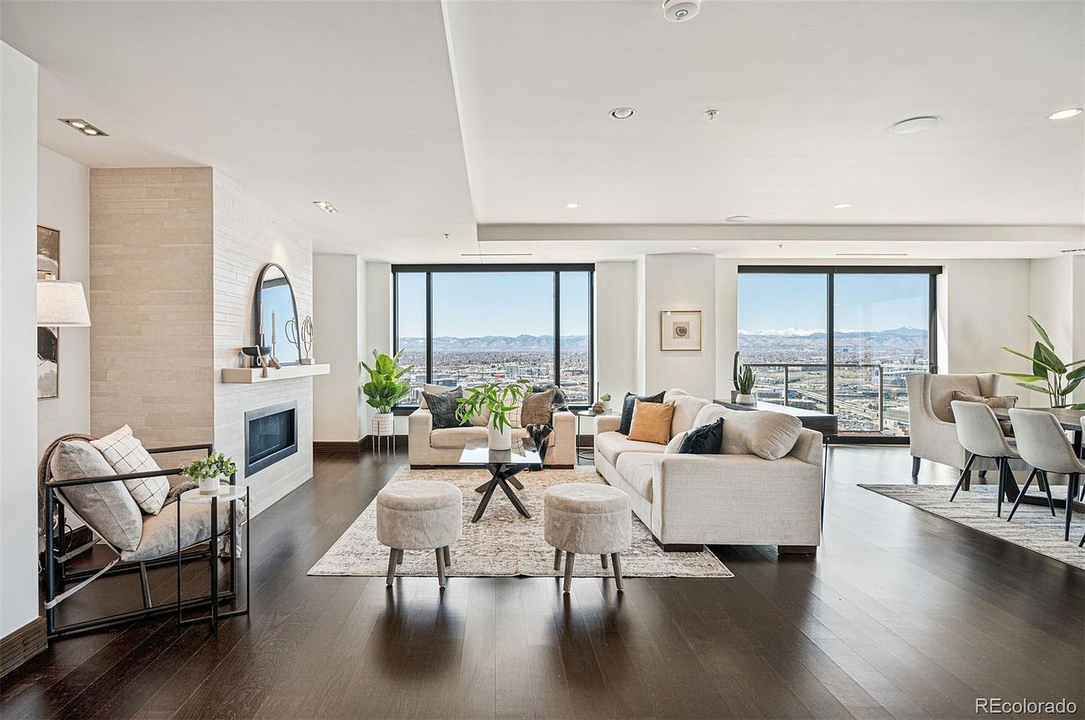 This is a bright and spacious living room featuring dark hardwood floors, a modern fireplace, and large windows offering a stunning city view. The room is furnished with comfortable seating, including a sofa, armchairs, and ottomans, arranged around a central coffee table. The overall impression is one of luxury and comfort, perfect for relaxing or entertaining.