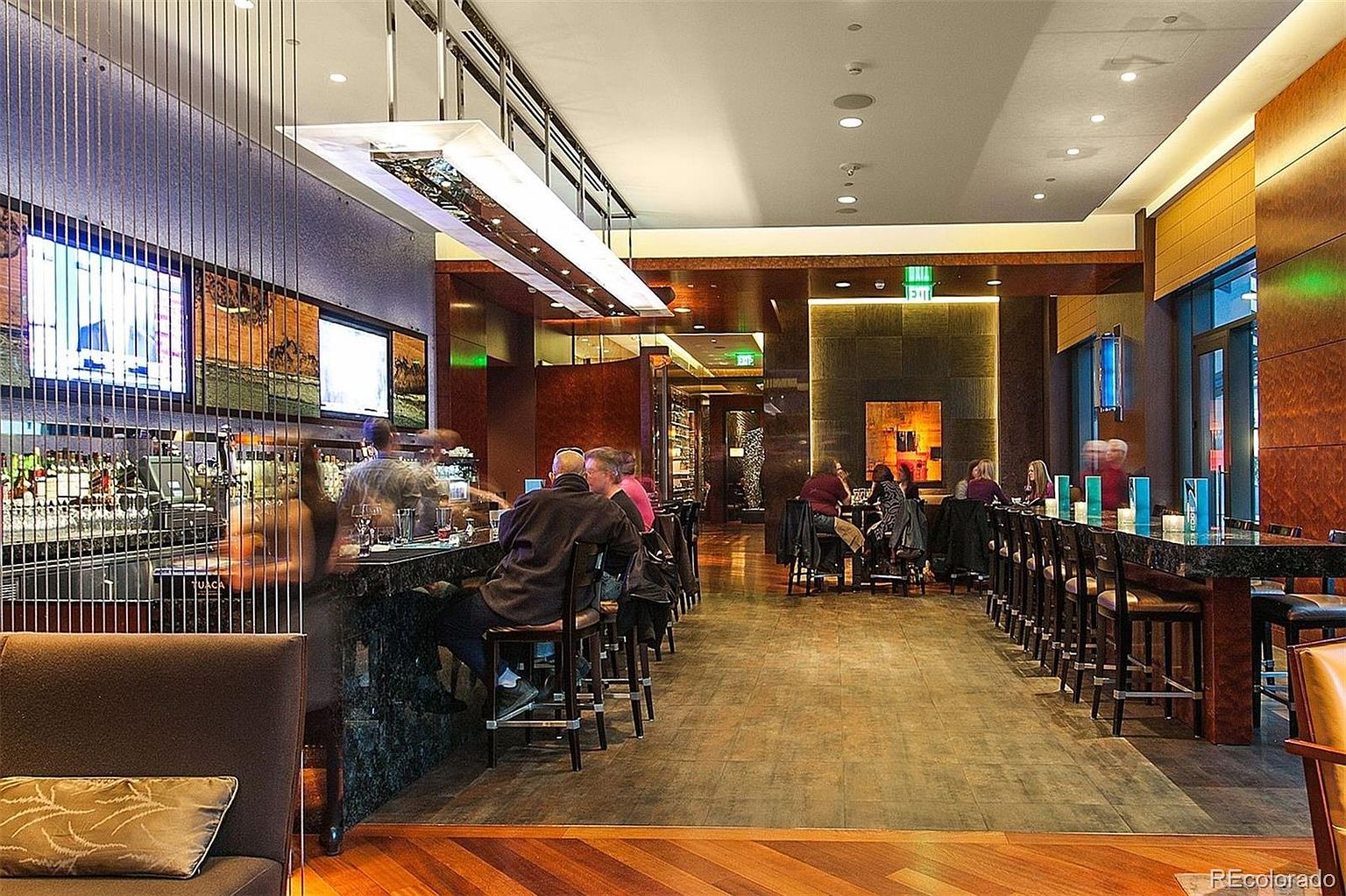 This interior shot showcases a stylish dining area within a restaurant or bar setting. The space features a long, dark marble bar with high-backed chairs, complemented by wooden floors and modern lighting fixtures. Patrons are seated at the bar and tables, creating a lively yet sophisticated atmosphere, suitable for a high-end dining experience.