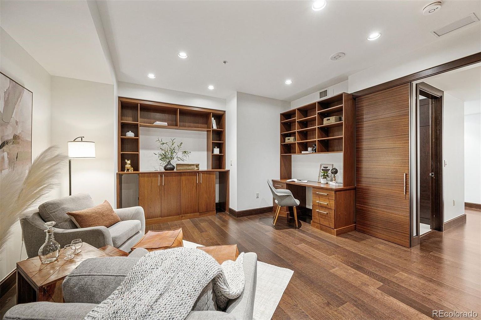 This interior shot showcases a well-appointed living room with built-in wooden shelving and cabinetry, complemented by a dedicated desk area. The room features hardwood flooring, comfortable seating arrangements, and a sliding wooden door, creating a warm and inviting atmosphere. The space is well-lit with recessed lighting, enhancing the overall appeal.