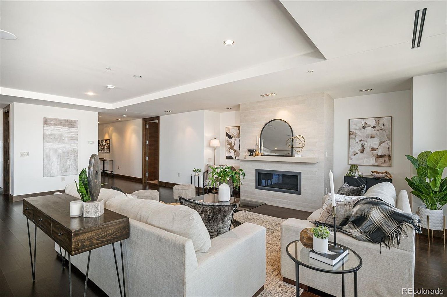 This is a well-lit living room featuring a modern fireplace with a decorative mirror above it. The room is furnished with comfortable-looking sofas, a dark wood console table, and a glass-topped coffee table. The dark wood flooring contrasts nicely with the light-colored walls and furniture, creating a sophisticated and inviting atmosphere.
