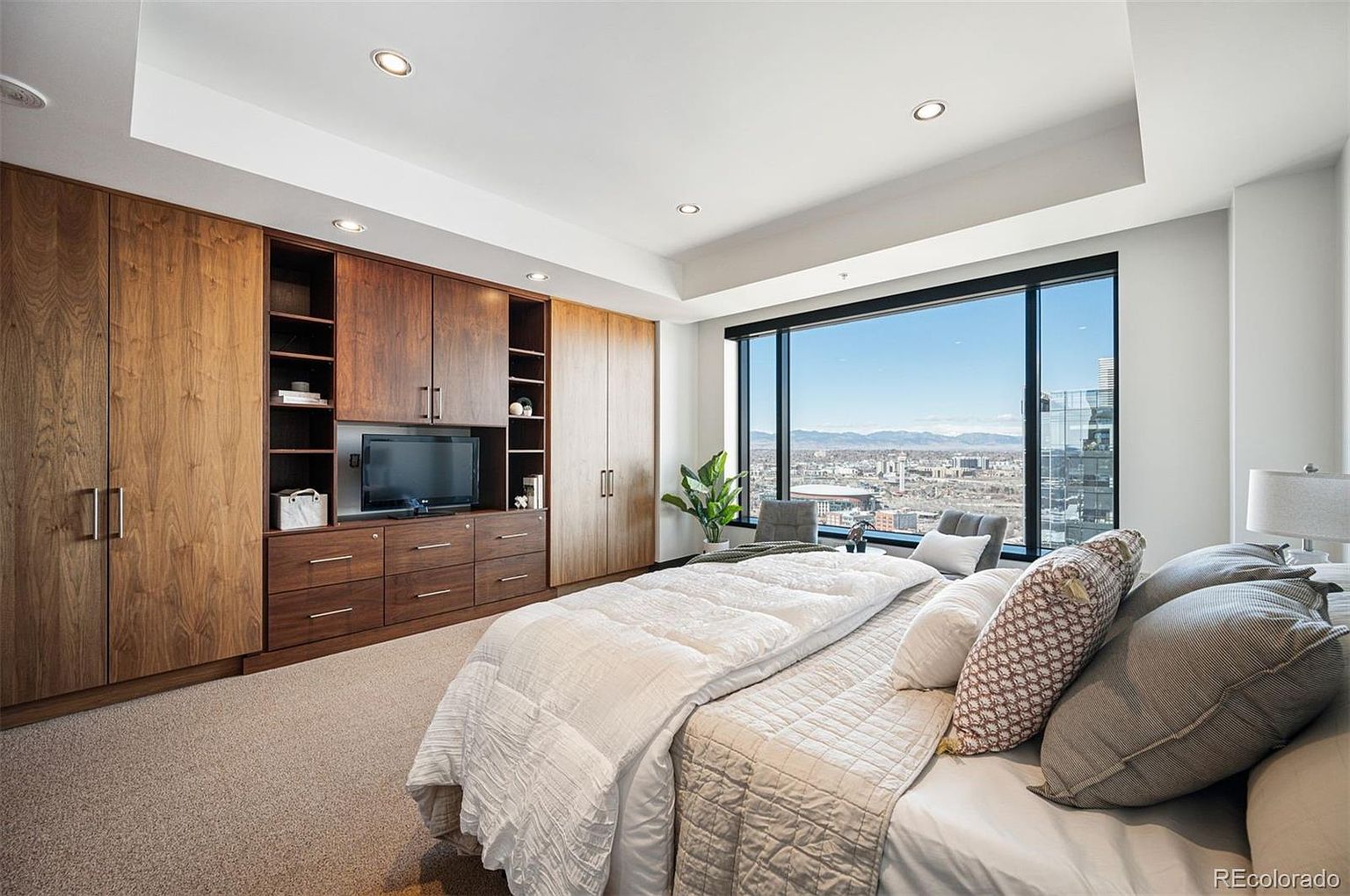 This is a primary bedroom featuring a large window with a city view, built-in wooden cabinetry with a television, and a king-size bed with white bedding and decorative pillows. The room has a neutral color palette and carpeted floors, creating a serene and luxurious atmosphere. The perspective is from the foot of the bed, looking towards the window and built-ins.