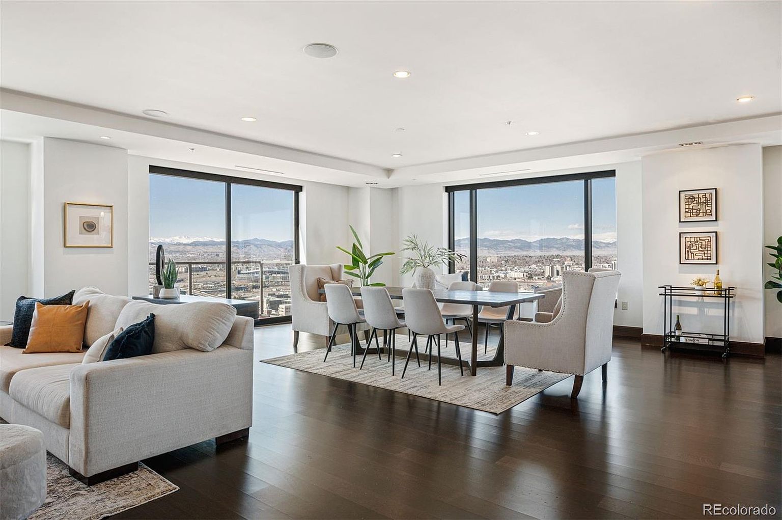 This is a spacious and modern living and dining area featuring dark hardwood floors and large windows with city and mountain views. The living room includes a comfortable beige sofa with decorative pillows, while the dining area has a sleek table with upholstered chairs and wingback chairs at the ends. The open layout and abundant natural light create an inviting and luxurious atmosphere.