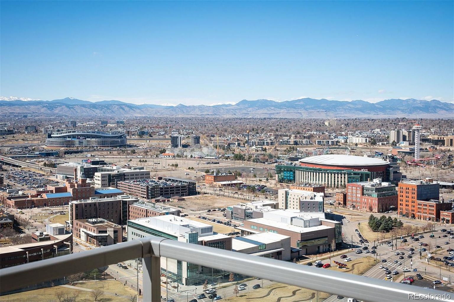 This aerial view showcases a vibrant cityscape with a mix of modern and traditional architecture. Several large stadiums and arenas are visible, surrounded by parking lots and roadways bustling with traffic. In the background, a stunning mountain range provides a scenic backdrop, enhancing the urban landscape.