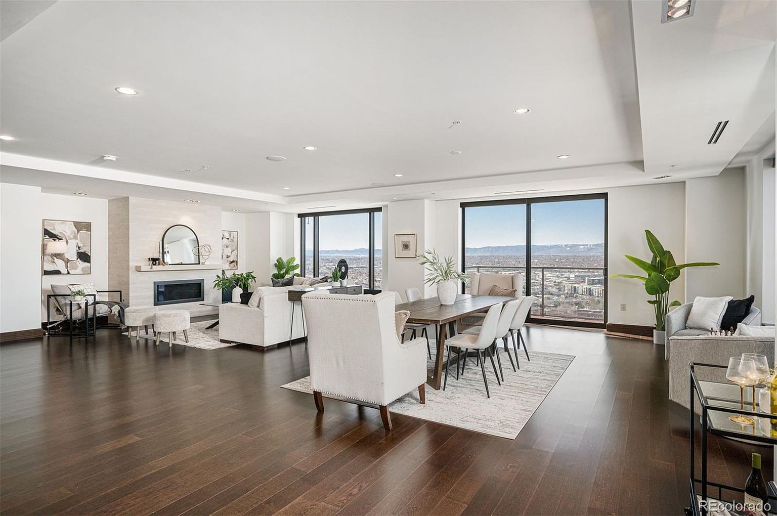 This is a spacious and luxurious living and dining area featuring dark hardwood floors, a modern fireplace, and large windows offering city views. The room is furnished with elegant seating, including sofas and dining chairs, and decorated with contemporary art and plants. The overall impression is one of sophistication and comfort, ideal for entertaining or relaxing.